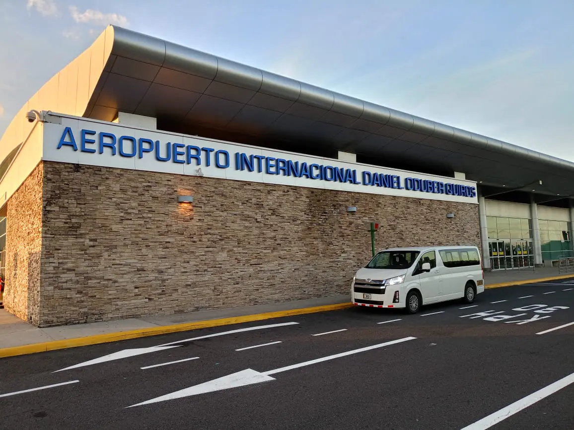 A white passenger van parked at the curb outside the Daniel Oduber Quirós International Airport terminal in Liberia